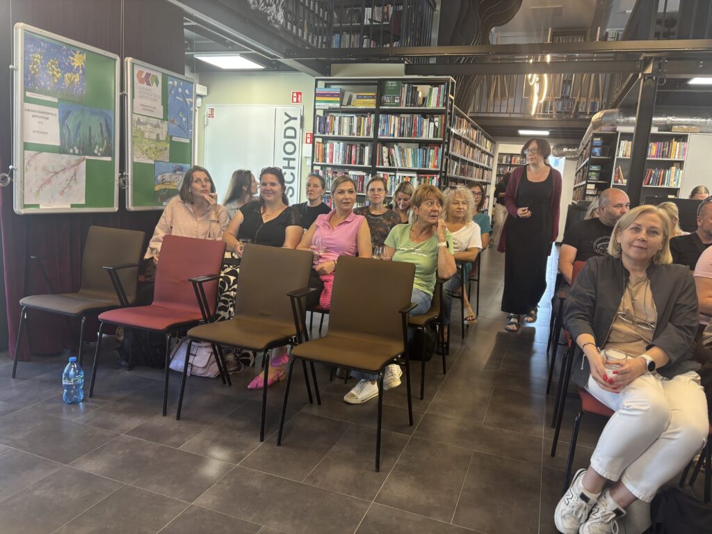 Sala spotkań w nowoczesnej bibliotece z regałami pełnymi książek w tle. Kilkanaście osób siedzi na krzesłach ustawionych w rzędach i oczekuje na rozpoczęcie wydarzenia. Po lewej stronie znajdują się plansze z pracami plastycznymi oraz baner z napisem „Lubuskie Konfrontacje Artystyczne”. W centralnej części widoczne są puste krzesła, a za nimi uczestnicy zgromadzeni między regałami. Wnętrze jest dobrze oświetlone, z ciemną podłogą i metalowymi konstrukcjami antresoli nad salą.