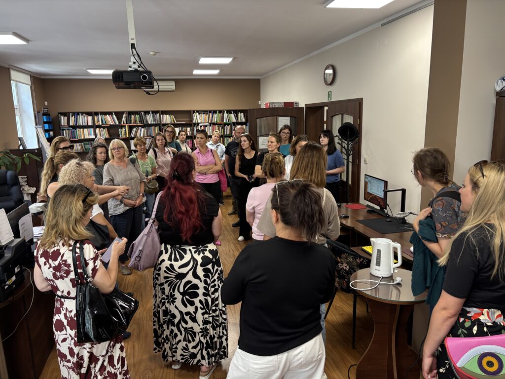 Pomieszczenie biblioteczne lub biurowe z regałami pełnymi książek w tle. W centralnej części stoi grupa około dwudziestu osób zgromadzonych wokół osoby prowadzącej prezentację lub oprowadzanie. Uczestnicy słuchają, stojąc między biurkami wyposażonymi w komputery i sprzęt biurowy. Nad salą zawieszony jest projektor, a na ścianie widoczny jest zegar. Wnętrze jest jasne, z drewnianą podłogą i dużą liczbą książek na półkach, co podkreśla biblioteczny charakter miejsca.