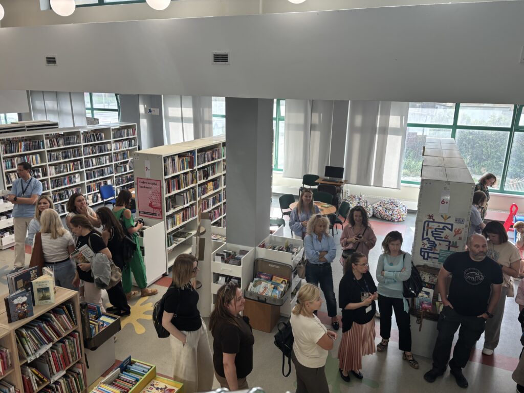 Widok z góry na przestrzeń biblioteczną z wysokimi regałami wypełnionymi książkami. Między półkami stoi i rozmawia grupa kilkunastu uczestników wydarzenia. W centralnej części znajdują się regały oznaczone kategoriami wiekowymi „6+” i „9+”, a na jednym z nich plakat z hasłem „Dobre książki kochamy”. W tle widoczne są duże okna z jasnymi roletami, stoliki, krzesła oraz kolorowe pufy. Jasne, przestronne wnętrze oświetlają naturalne światło i okrągłe lampy sufitowe.