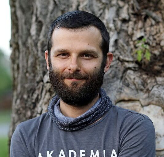Portret lekko uśmiechniętego mężczyzny stojącego przy pniu drzewa. Ma krótkie ciemne włosy i brodę, ubrany jest w szarą koszulkę oraz ciemną chustę na szyi.