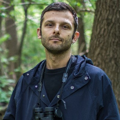 Portret mężczyzny stojącego w lesie. Ma krótkie ciemne włosy, ubrany jest w granatową kurtkę, a na szyi ma zawieszoną lornetkę. W tle widoczne są drzewa i zielona roślinność.
