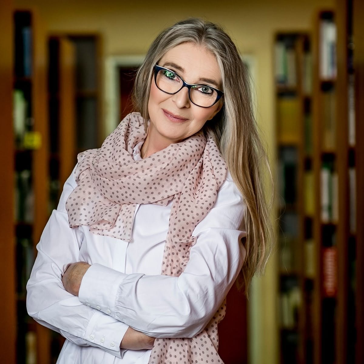 Violetta Nowakowska, w tle książki, kobieta trzyma się pod boki, ma szal i białą koszulę, długie rozpuszczone włosy i okulary. 