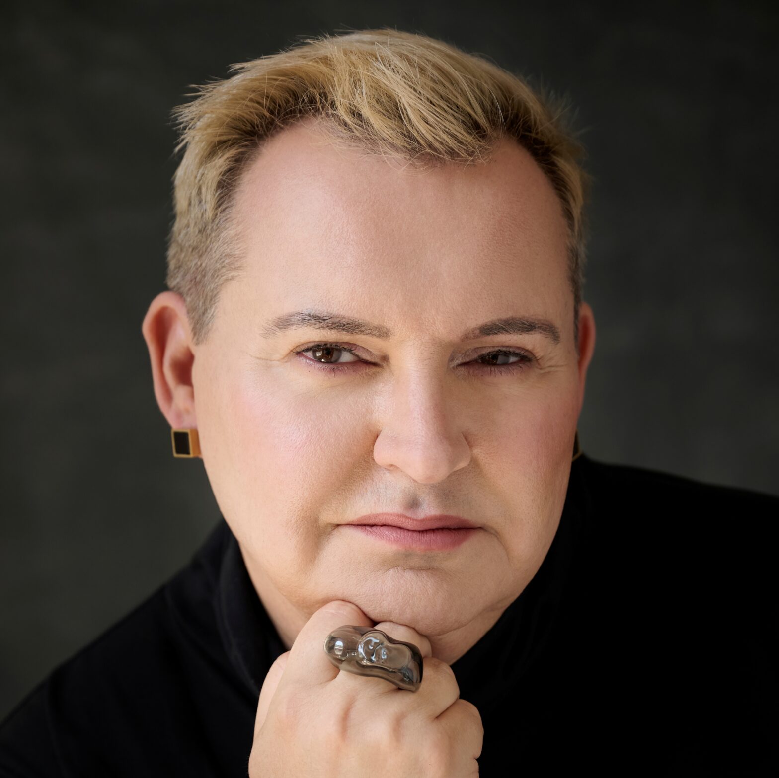Michał Witkowski, Portret osoby o krótkich, jasnobrązowych włosach, ubranej w czarną odzież. Osoba patrzy prosto w obiektyw z lekko zmrużonymi oczami, opierając brodę na dłoni. Na palcu dłoni ma dużą, srebrzystą rzeźbiarską obrączkę, a w uszach geometryczne, czarne kolczyki. Tło jest ciemne i jednolite.