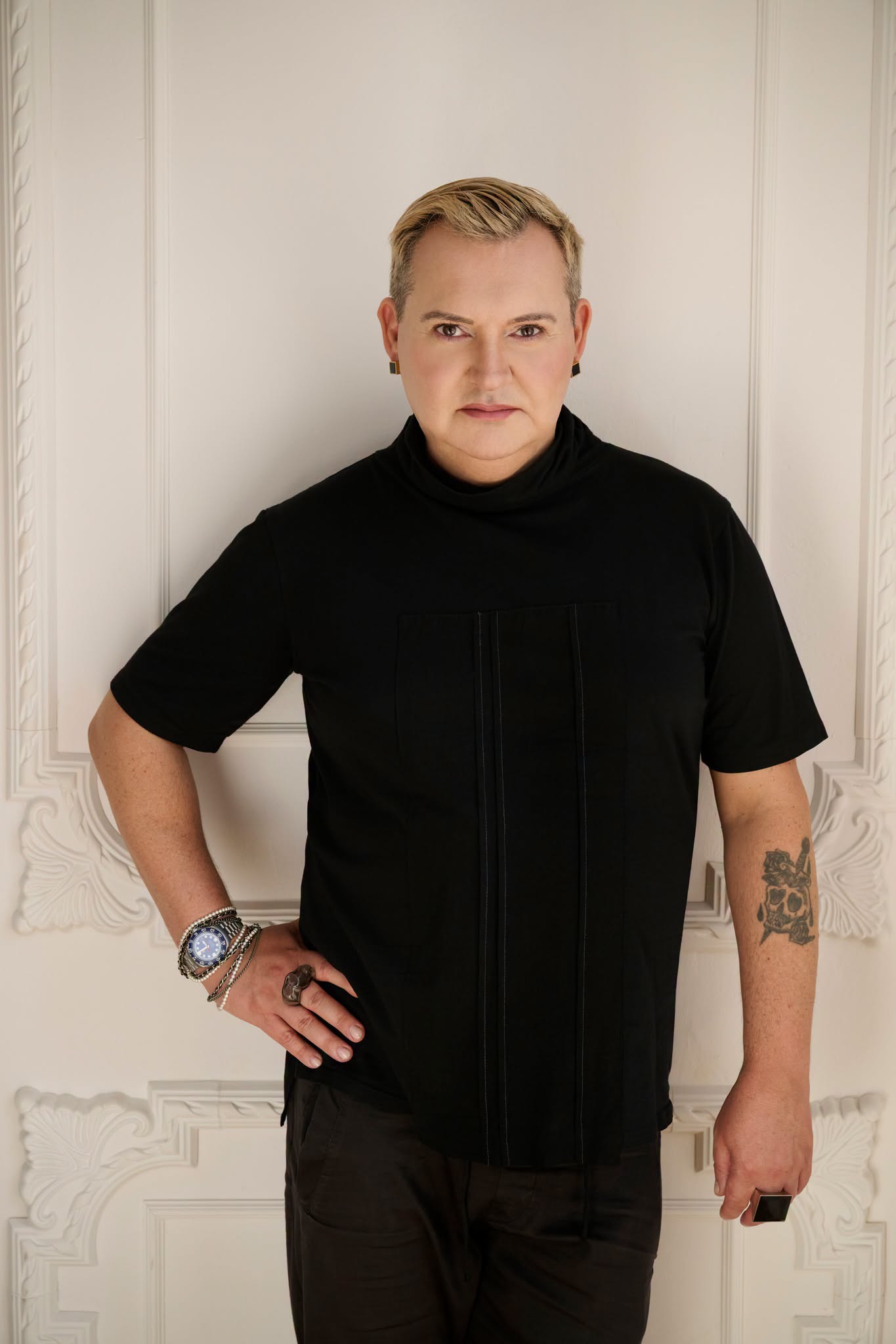 Michał Witkowski, stoi na tle białych ozdobnych drzwi, jest ubrany na czarno, prawa ręka na biodrze, patrzy w obiektyw,