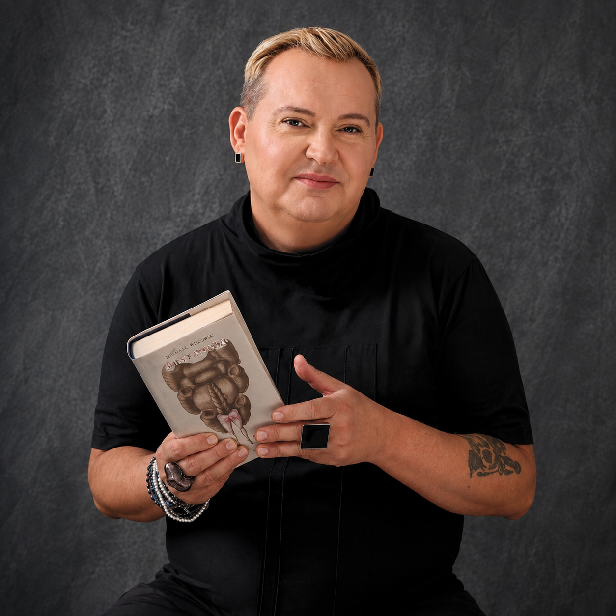 Michał Witkowski w czarnym golfie trzyma swoją nową książkę. Ma tatuaż i pierścienie na rękach, blond włosy, szare tło.