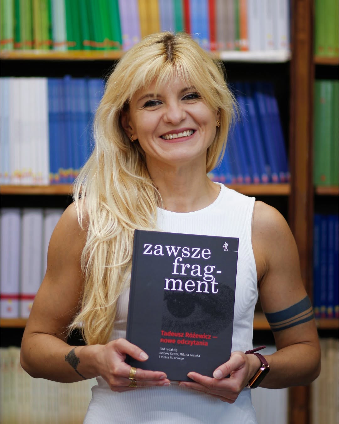 Justyna Kowal ubrana w białą bluzkę na tle regału z książkami, w rękach trzyma swoją publikację