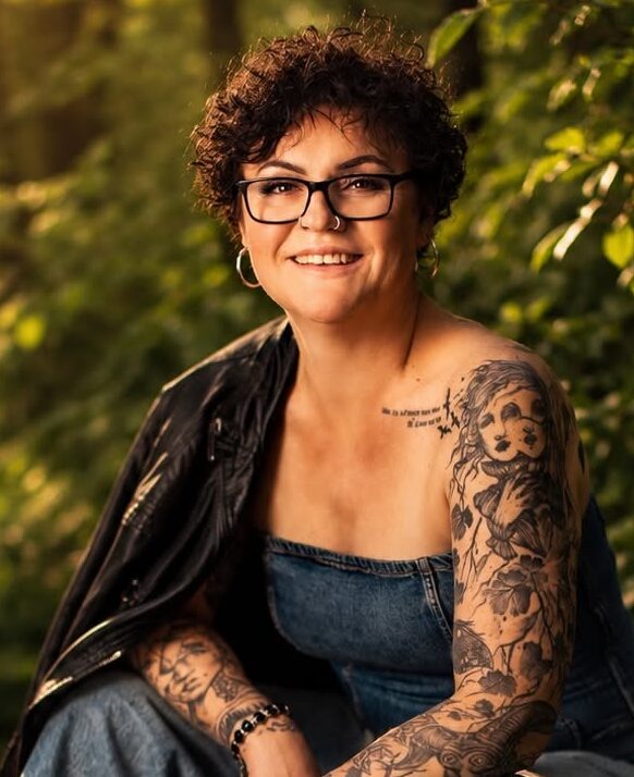 Portret uśmiechniętej kobiety w krótkich, kręconych, ciemnobrązowych włosach. Nosi okulary w czarnej oprawce, ma kolczyki kółka w nosie i uszach, wytatuowane ręce, przez ramię ma przewieszoną czarną kurtkę, ubrana w dżinsowy kombinezon, na tle zielonych liści.