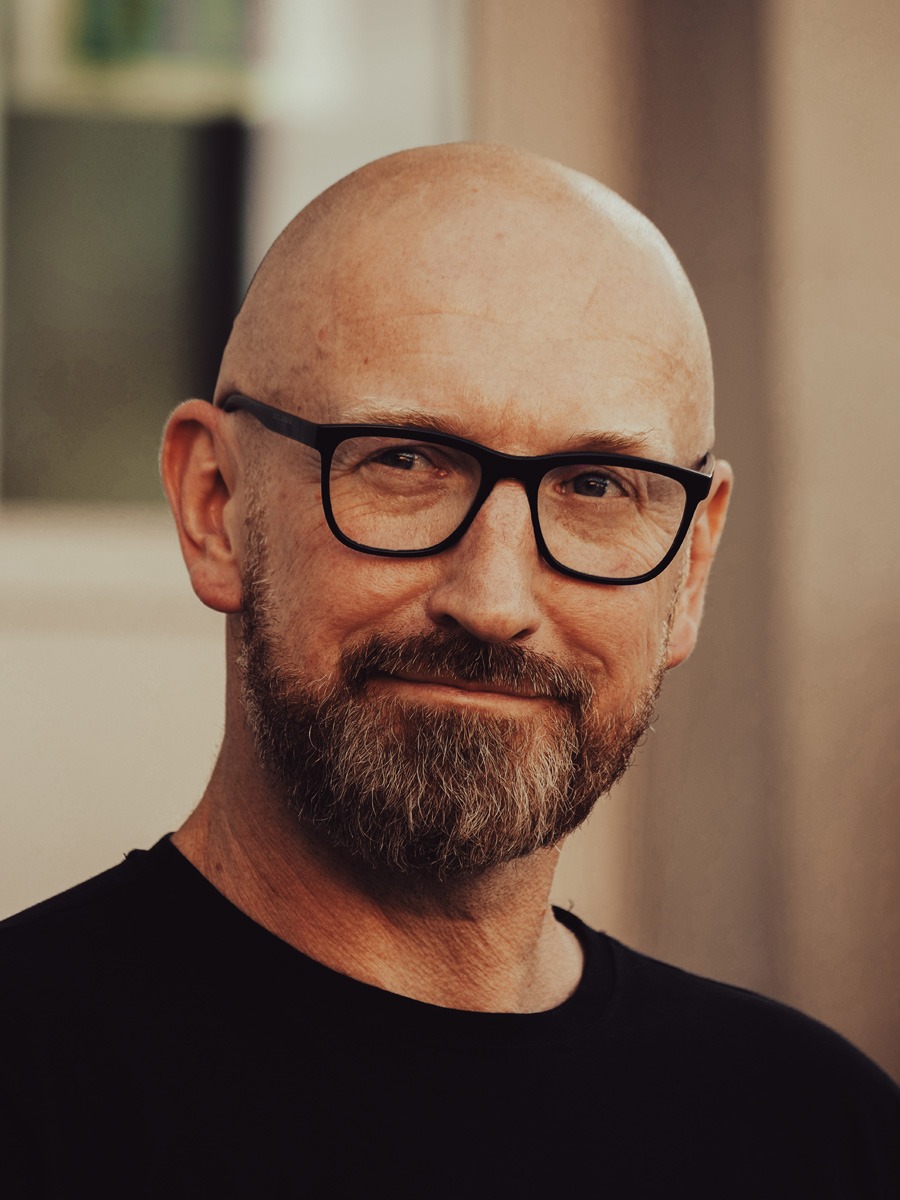 Portret łysego mężczyzny z krótką brodą w okularach z czarną oprawką, patrzy w bok obiektywu, ubrany w czarną koszulkę, na beżowym tle.