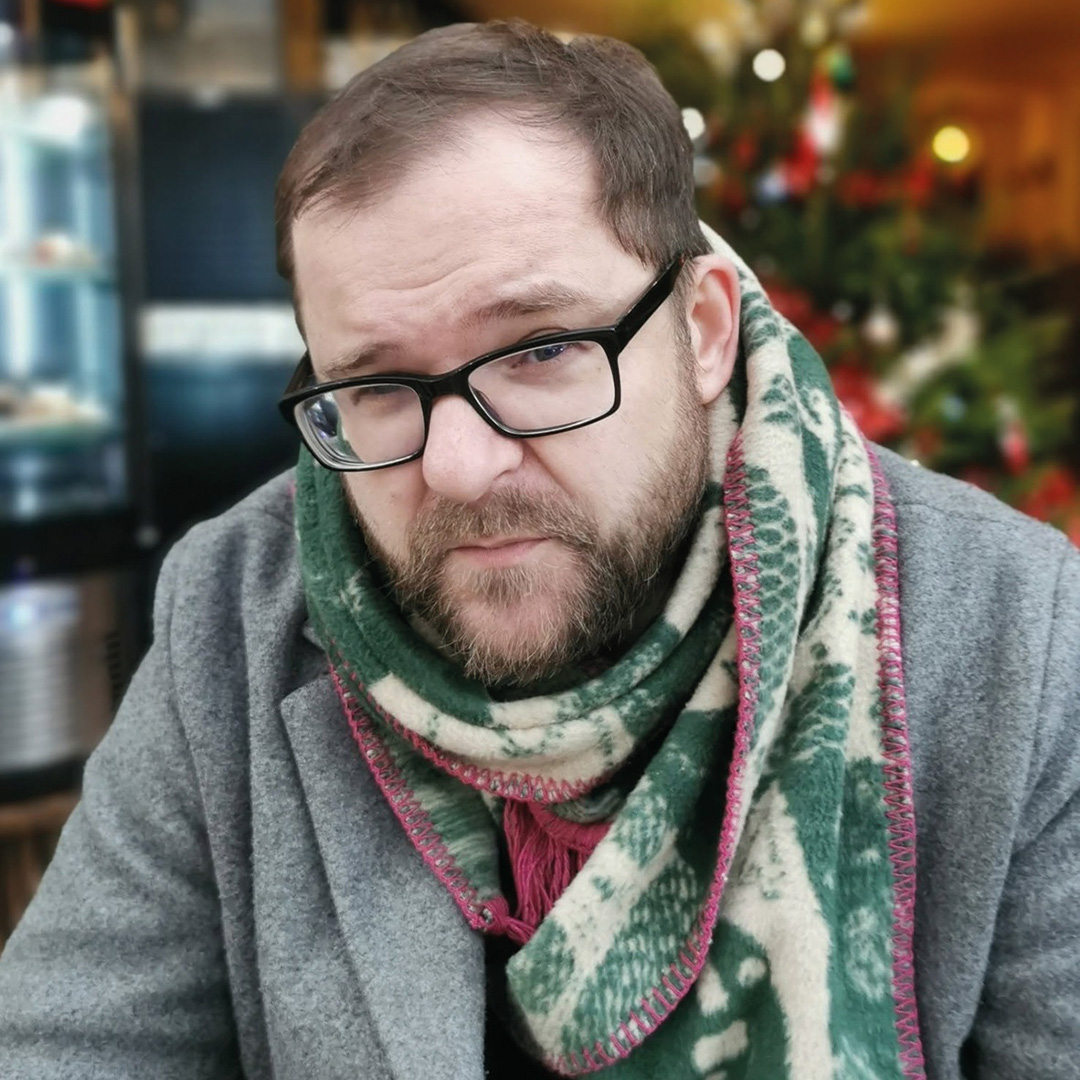 Portret osoby w okularach siedzącej we wnętrzu lokalu. Osoba ma na sobie szary płaszcz oraz gruby zielono-biały szalik. W tle widoczne są rozmyte dekoracje i świąteczne światełka.