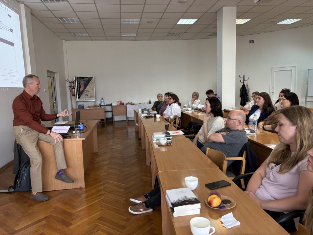 W przestronnej sali szkoleniowej osoba prowadząca prezentację siedzi na brzegu biurka obok laptopa i ekranu projekcyjnego, zwracając się do kilkunastu uczestników siedzących przy ustawionych w podkowę stołach. Na stołach znajdują się książki, notatniki, kubki z napojami, telefon oraz przekąski. W tle widać stolik z poczęstunkiem, wieszak na odzież i jasne ściany sali.
