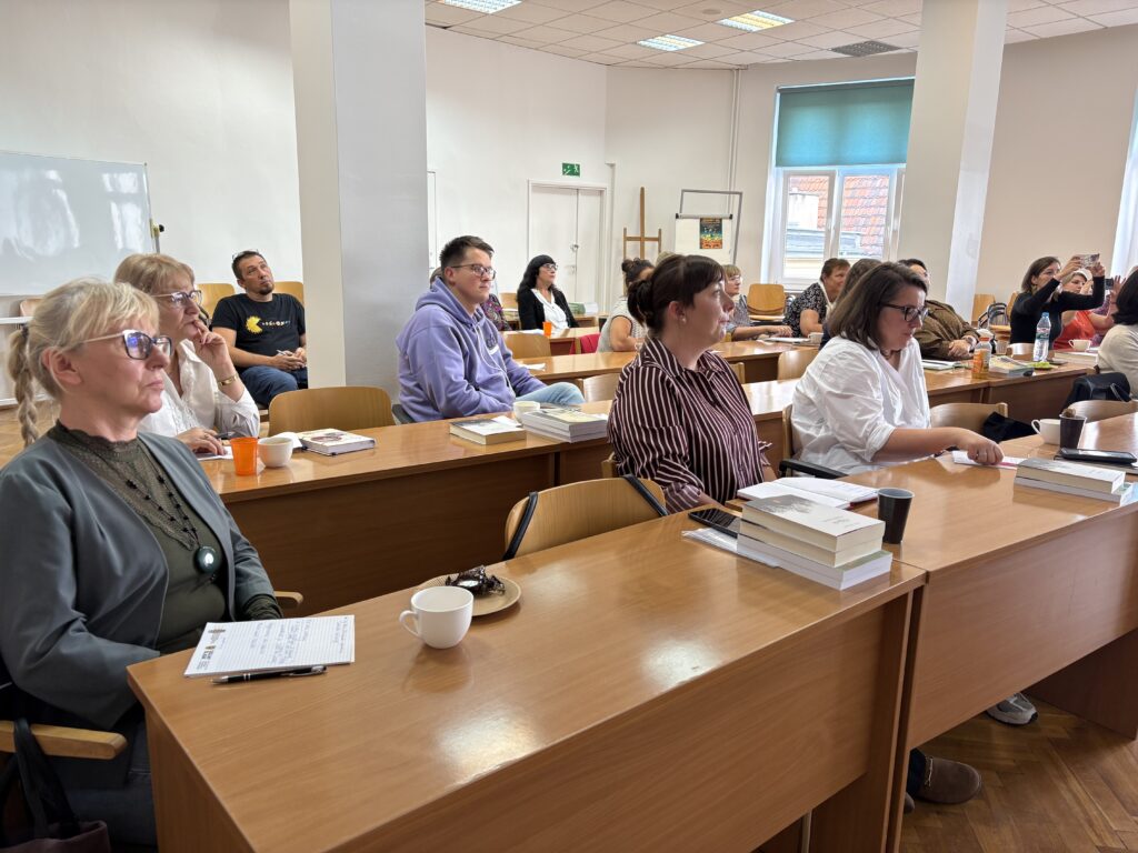 W jasnej sali szkoleniowej kilkanaście osób siedzi przy drewnianych stołach ustawionych w rzędach. Na stołach znajdują się książki, notatniki, kubki z napojami, telefony oraz drobne przekąski. Uczestnicy są zwróceni w stronę prezentacji odbywającej się poza kadrem po lewej stronie. W tle widoczne są białe ściany, kolumny, tablica suchościeralna oraz duże okno z niebieską roletą. Sala jest dobrze oświetlona światłem dziennym i oświetleniem sufitowym.