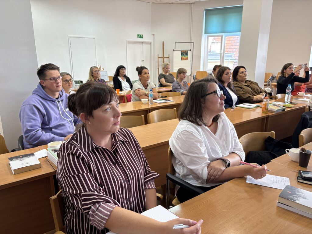 W jasnej sali szkoleniowej grupa uczestników siedzi przy drewnianych stołach ustawionych w kilku rzędach. Na pierwszym planie widoczne są osoby z notatnikami i długopisami, a na stołach znajdują się książki, kubki, butelki z napojami oraz telefony. Wszyscy są zwróceni w stronę prezentacji prowadzonej poza kadrem po lewej stronie. W tle znajdują się wysokie okna z niebieską roletą, białe ściany, drzwi oraz dodatkowe krzesła ustawione pod ścianą. Sala jest dobrze oświetlona światłem dziennym i oświetleniem sufitowym.