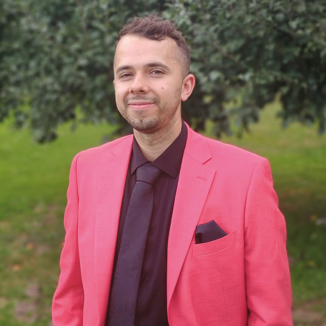 Portret osoby ubranej w różową marynarkę, czarną koszulę i czarny krawat. Zdjęcie wykonano na świeżym powietrzu, na tle zielonych drzew.