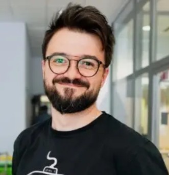 Portret osoby w okularach i z zarostem, ubranej w czarną koszulkę. Zdjęcie wykonano we wnętrzu z przeszklonymi ścianami i jasnym tłem.