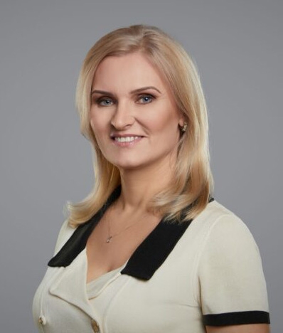 Portret kobiety o blond włosach do ramion, ubranej w jasną bluzkę z czarnym kołnierzykiem i czarnymi lamówkami przy rękawach. Kobieta uśmiecha się i patrzy w obiektyw. Zdjęcie wykonano na jednolitym szarym tle.