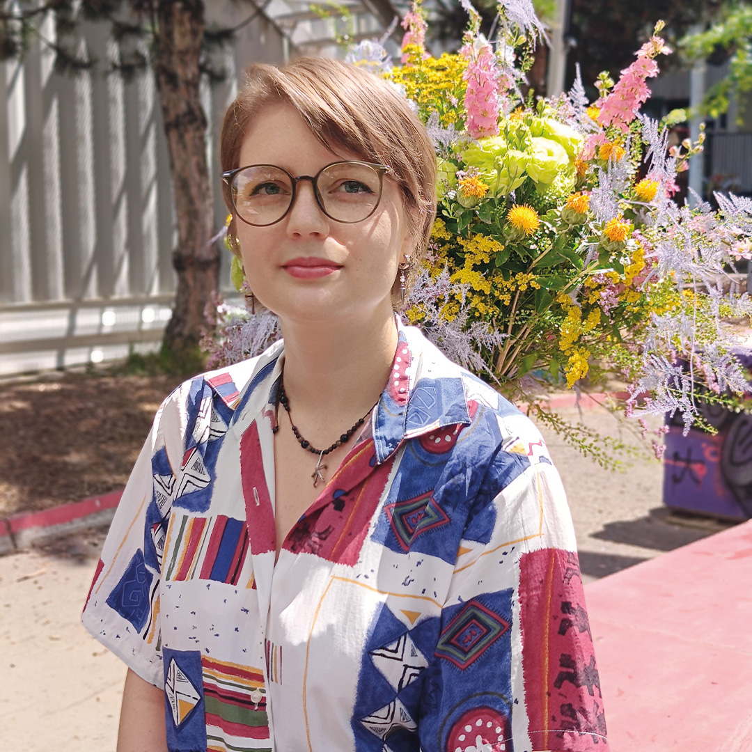 Portret osoby w okularach, siedzącej na zewnątrz na tle dużego, kolorowego bukietu kwiatów. Osoba ma na sobie wzorzystą koszulę w odcieniach niebieskiego, czerwieni i bieli.