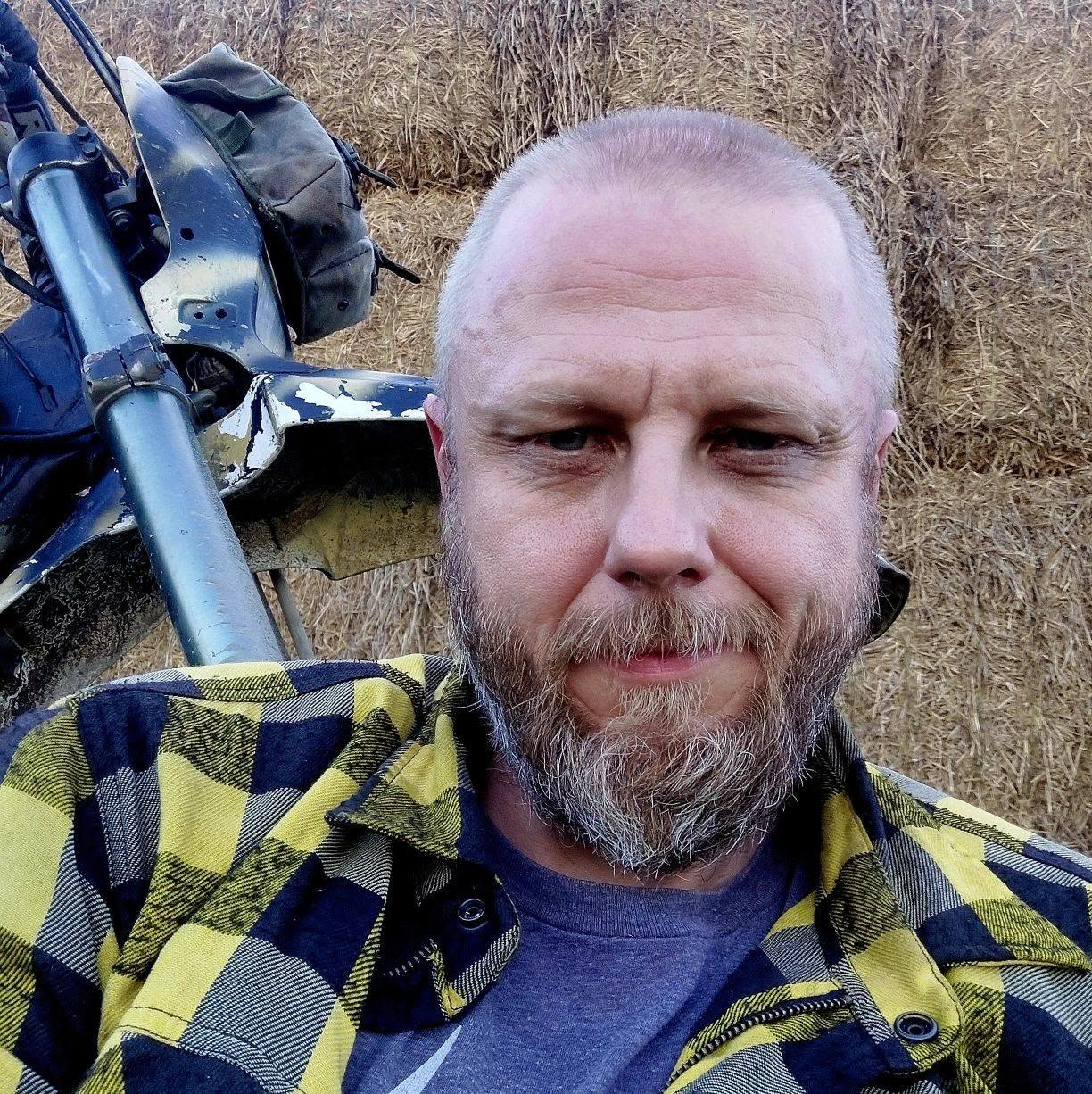 Portret osoby z brodą, ubranej w żółto-czarną koszulę w kratę. W tle widoczne są bele siana oraz fragment motocykla lub roweru terenowego.
