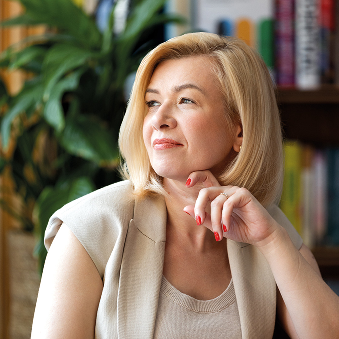 Portret osoby siedzącej przy stole w bibliotece. W tle widoczne są regały z książkami i duża zielona roślina. Osoba opiera brodę na dłoni i patrzy w bok.