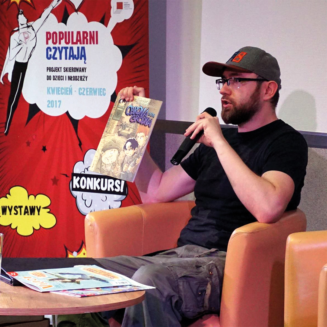 Osoba siedzi w fotelu podczas spotkania autorskiego i trzyma w dłoni komiks. W drugiej dłoni trzyma mikrofon. W tle widoczny jest kolorowy baner promujący projekt czytelniczy.