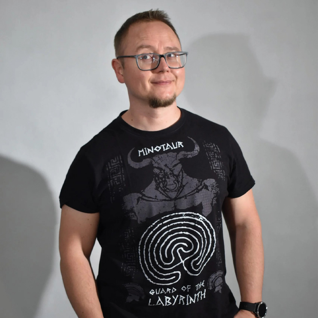 Portret osoby w okularach stojącej na jednolitym, jasnym tle. Osoba ma na sobie czarny T-shirt z dużą grafiką labiryntu i minotaura oraz zegarek na nadgarstku.