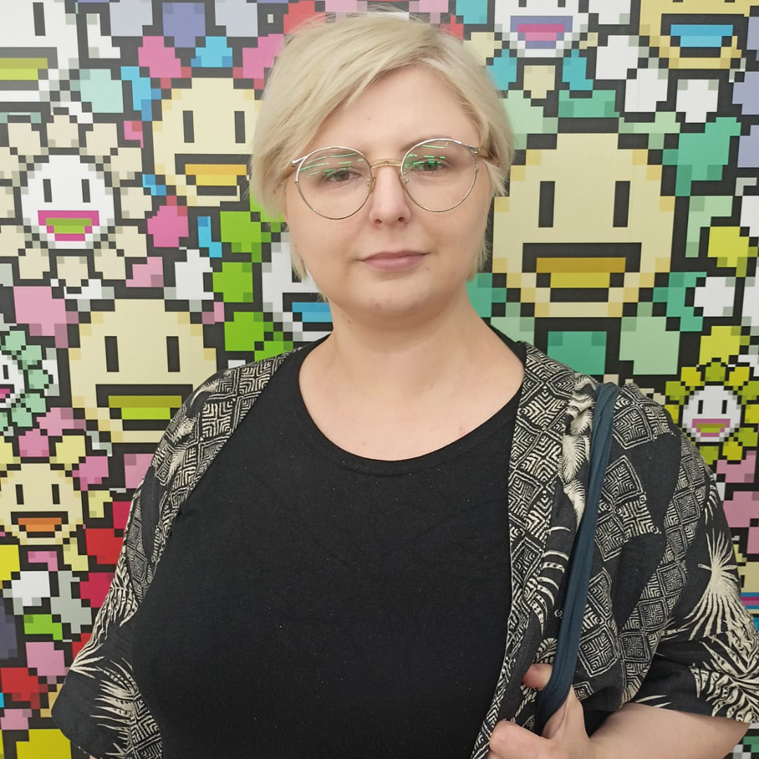 Portret osoby w okularach stojącej na tle kolorowej grafiki w stylu pikselowym. Osoba ma krótkie jasne włosy i ubrana jest w czarną bluzkę oraz wzorzyste okrycie.