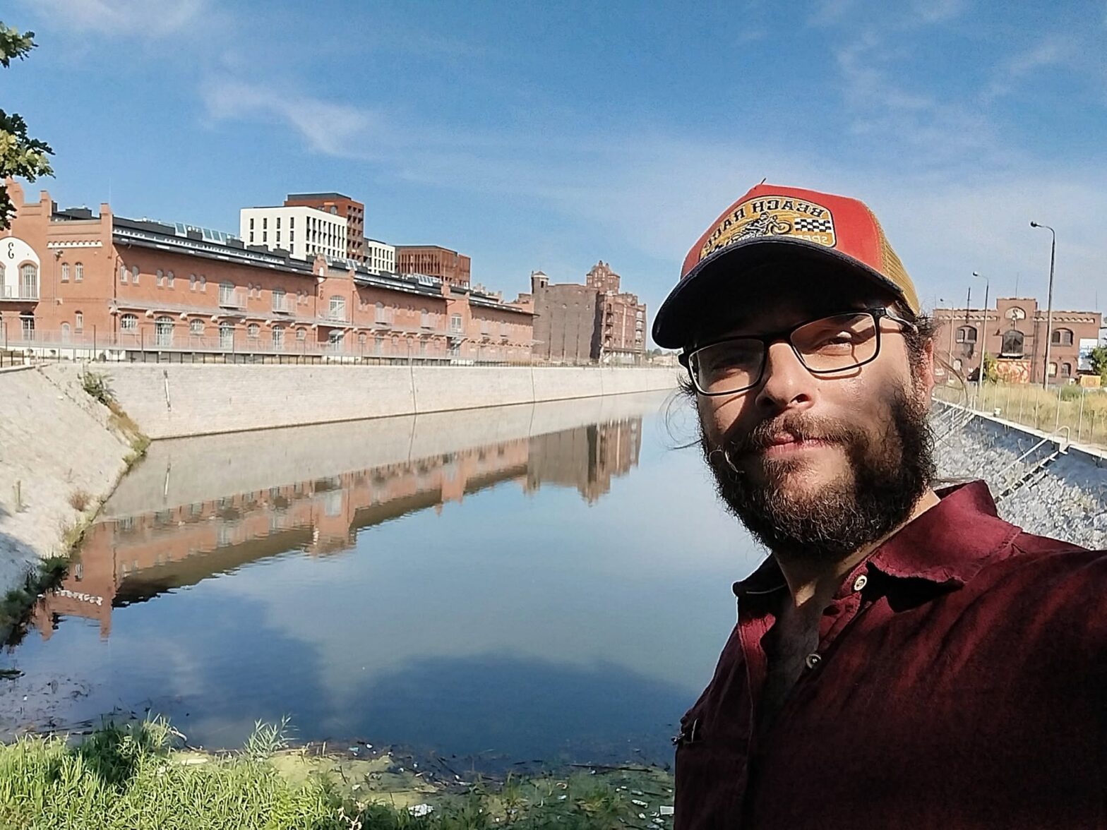 Selfie osoby w czapce i okularach nad kanałem wodnym. W tle ceglane budynki industrialne odbijające się w spokojnej wodzie pod błękitnym niebem.