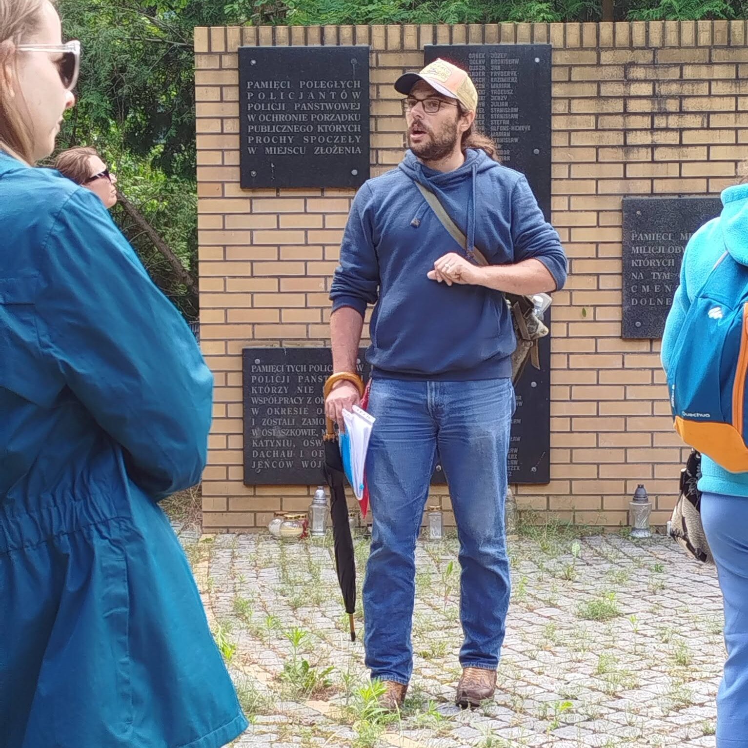 Osoba w czapce i okularach prowadzi oprowadzanie przy memoriale z tablicami pamiątkowymi. Wokół stoją uczestnicy, a w tle widoczny jest ceglany mur i drzewa.